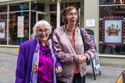 Foto von zwei älteren, eng beieinander stehenden Personen, beide lächeln. Die linke Person schaut dabei in die Kamera, die rechte in die Ferne. Beide Personen tragen Brillen und leichte Jacken. Im Hintergrund ist ein Geschäft mit beleuchteten Schildern zu sehen.