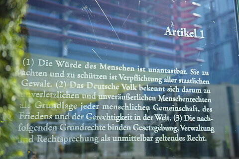 Artikel 1 des Grundgesetzes in weißer Schrift auf einer Glasscheibe vor einem Gebäude des Bundestags.